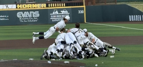 Baseball players in white uniforms pile on top of each other in a celebratory heap on an infield, with one player midair landing on the group. Several water bottles are scattered nearby on the grass and dirt. In the background, a green outfield wall displays advertisements and a “330” distance marker.