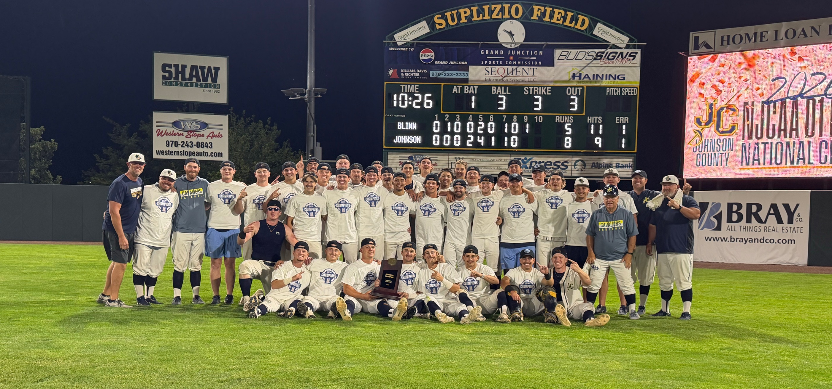 Large group of baseball players and staff pose on a grassy field at night in front of a scoreboard labeled “Suplizio Field.” The group wears matching light-colored “Champions” shirts; some people kneel in the front row while others stand behind them with arms around each other. A player in the center front holds a trophy. The scoreboard above shows a completed game with final runs of 8–5, along with inning-by-inning scoring. Bright stadium lights illuminate the scene, and advertising signs and a large digital display board are visible in the background.