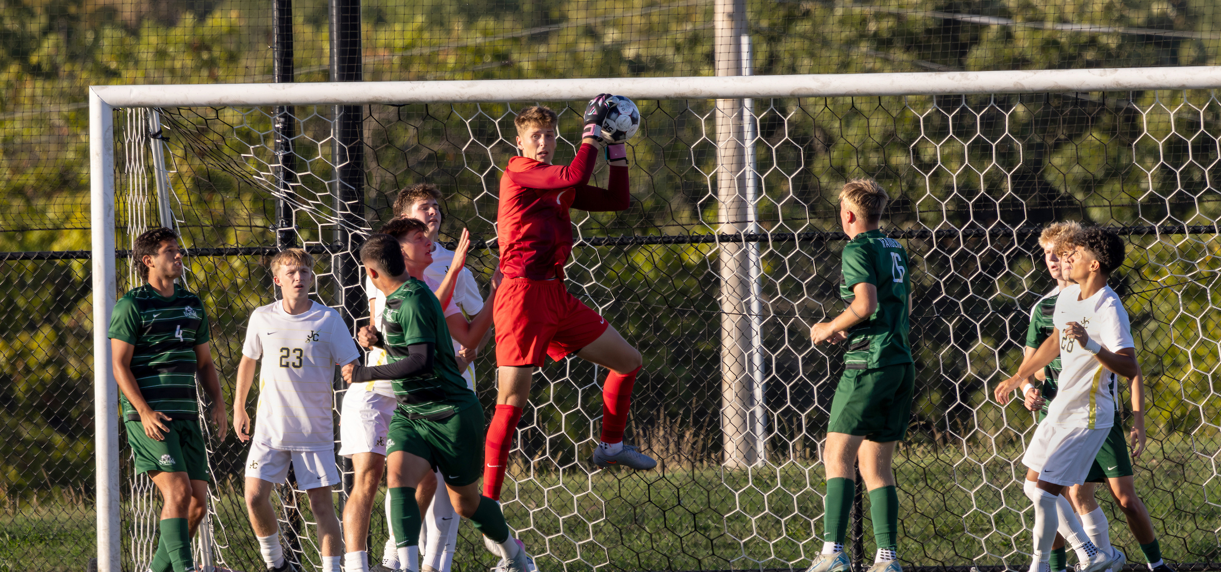The photo captures an intense soccer moment near the goal. A goalkeeper dressed in a bright red uniform is airborne, holding the ball securely with both hands just above the goal line. Several players from two different teams, wearing green and white uniforms, are positioned close to the goal, likely anticipating or reacting to the play. The goal net with hexagonal patterns is clearly visible, and the scene is set outdoors on a sunny day with green foliage in the background. The players appear to be in a defensive or attacking formation during what looks like a corner kick or a cross into the box.