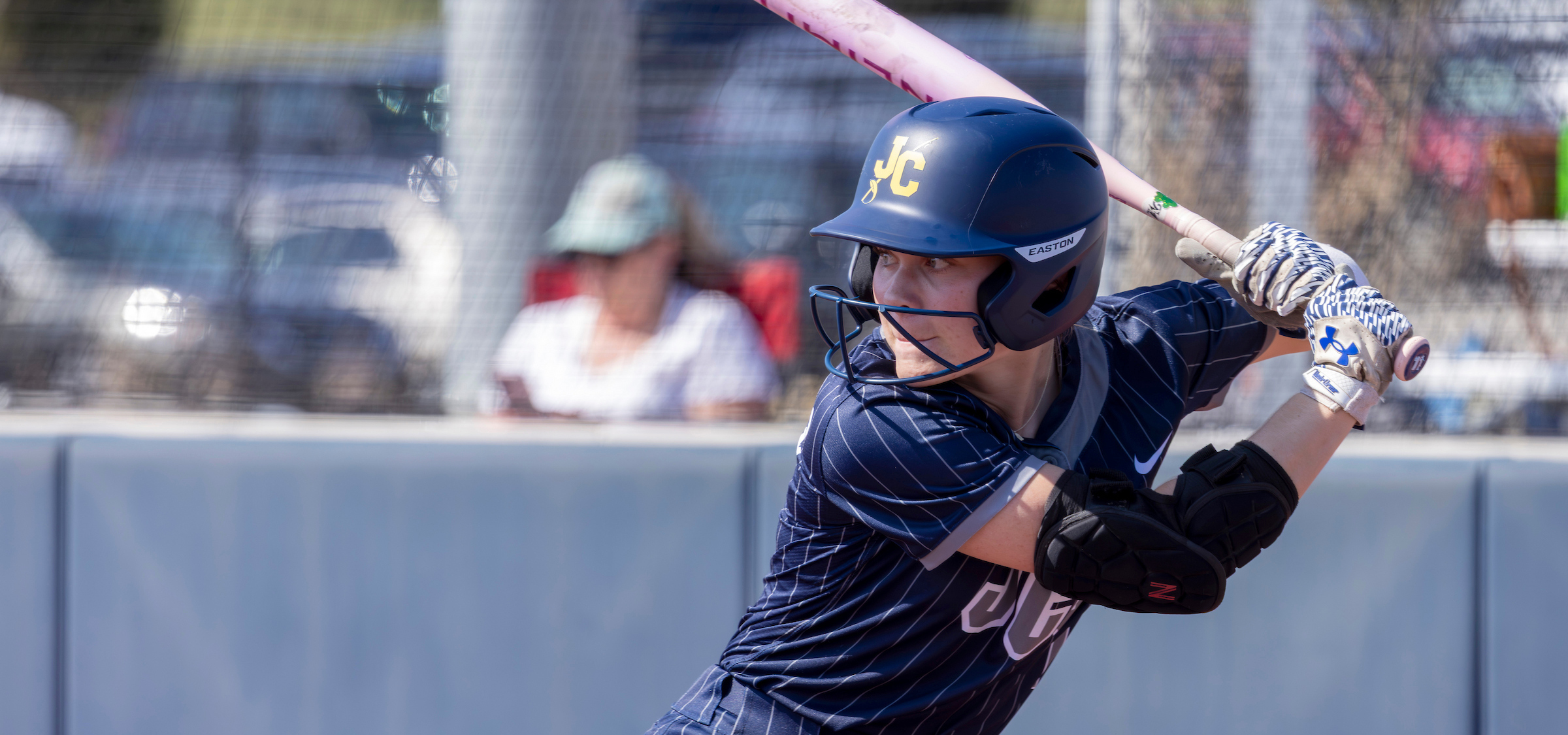 The image shows a baseball or softball player in the middle of a swing during a game. Here’s a detailed description:


Foreground:
The athlete is wearing a navy-blue uniform with thin white pinstripes and protective gear, including batting gloves, an elbow guard, and a helmet marked with stylized initials that appear to be “JC” in yellow. The player is gripping a pink bat and is captured at the moment of winding up or beginning a swing.


Action & Positioning:
The athlete’s stance is athletic and focused, with the upper body turned slightly and the front shoulder angled toward the pitcher’s direction. The player’s arms are extended, holding the bat diagonally across the frame. The scene conveys motion and game intensity.


Background:
Behind the player is a blurred outdoor setting that appears to be a sports complex. A chain-link fence and spectators seated in the background are visible. Beyond them are parked cars and bright daylight conditions, suggesting a warm, sunny day.


Colors & Details:
The image features mostly cool tones—navy blue and gray—contrasted by the bright pink bat. The lighting is natural, and the overall scene is sharp and action-focused.