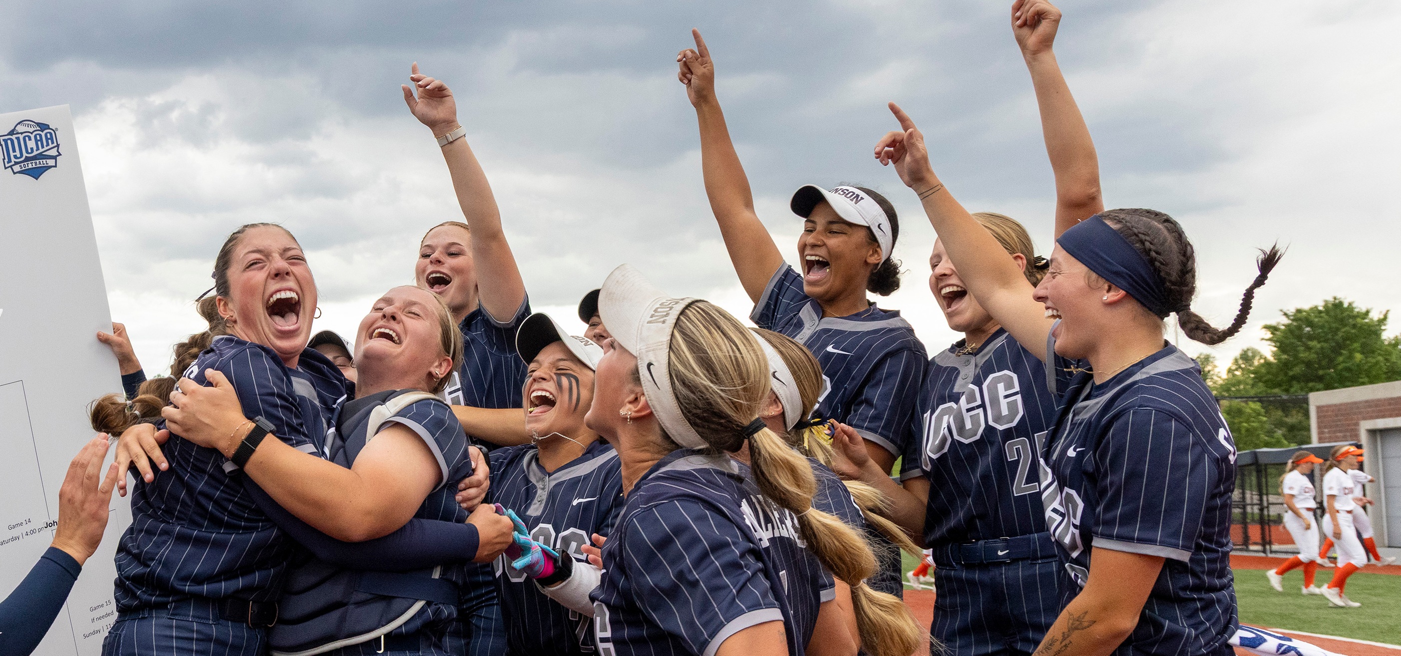 A group of softball players in dark blue pinstriped uniforms gather closely on a field, raising one arm upward and pointing while holding and cheering around a large white sign with a visible “NJCAA Softball” logo on the left. Several players wear visors or headbands, and some have eye black stripes. One player holds a colorful glove near the center of the group. In the background, a softball stadium setting includes green turf, a fence, a building, and another team in white and orange uniforms walking along the right side under a cloudy sky.
