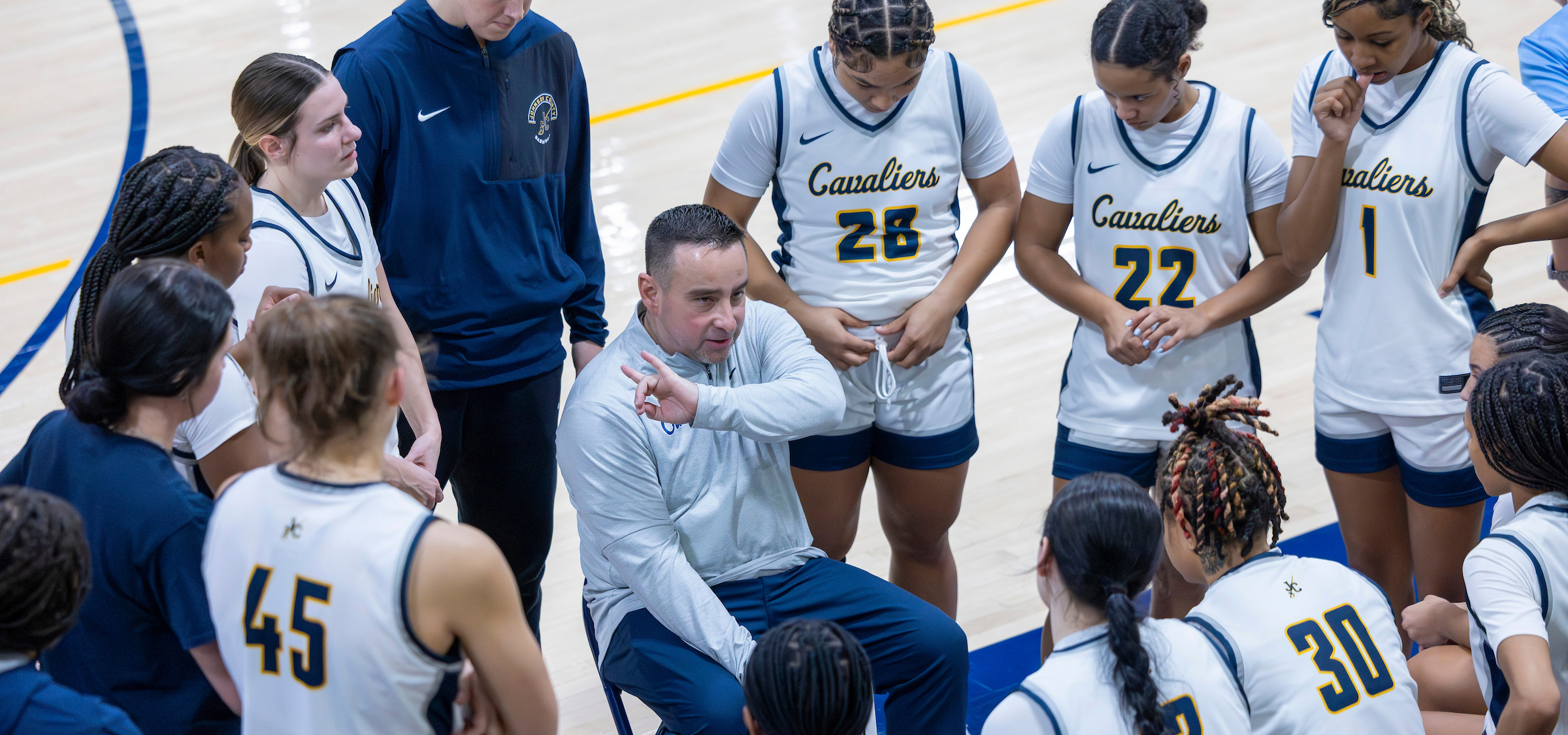 A basketball coach sits on a folding chair in the middle of a team huddle on an indoor court, gesturing with one hand while giving instructions. Players wearing white “Cavaliers” uniforms with navy and gold accents—numbers 28, 22, 30, and 45 visible—stand in a circle around the coach, listening closely. An additional staff member in a navy jacket stands nearby. The hardwood floor and painted court lines are visible beneath the group, with all attention focused inward on the strategy discussion.