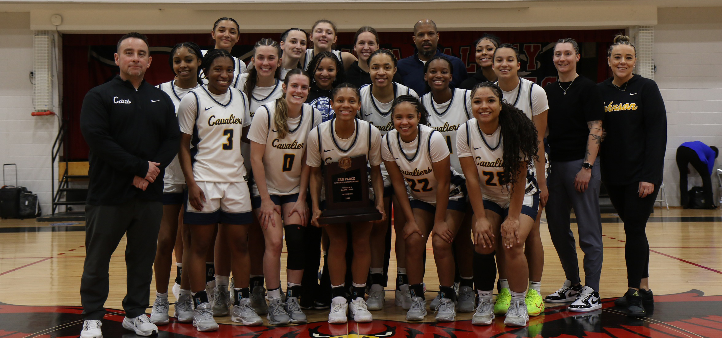 The image shows a large group of basketball team members and coaching staff gathered together on a gymnasium court for a posed team photo. Here’s a detailed description:
Overall Setting

The group is standing on a hardwood basketball court.
A red-and-black gym logo is visible beneath their feet.
The background includes gym walls, padding, and a partially visible banner behind the group.

Group Composition

The group consists of players wearing matching basketball uniforms and several staff members standing on each side.
The players are wearing light-colored jerseys with dark trim. The word “Cavaliers” is printed across the front of the uniforms.
Each uniform includes a visible number, such as 3, 0, 22, and others.
Team members are arranged in multiple rows: some standing in back, some slightly crouched or leaning forward in front.

Objects and Details

A central person in the front row is holding a trophy/plaque, which appears to signify a tournament placement or championship achievement.
Footwear varies, with several kinds of athletic shoes in different colors, including white, black, and neon yellow.
One staff member on the left is wearing a dark athletic outfit with the word “Coach” on the chest.
On the right side, another staff member is dressed in athletic apparel with the word “Cavaliers” on the shirt.

Lighting and Atmosphere

The lighting is typical of an indoor gym, evenly illuminating the group.
The overall setting suggests a celebratory moment—likely after a tournament game or award ceremony.