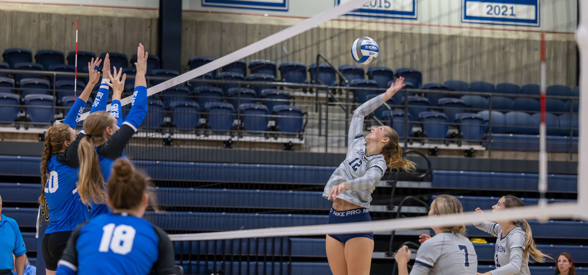 The photo captures an intense moment during a volleyball match:

A player wearing a gray jersey with the number 12 is airborne, attempting to spike the ball over the net.
On the opposing side, three players in blue jerseys are jumping together to block the shot.
The setting is a gymnasium, with empty bleachers in the background.
Banners on the wall display the years 2015 and 2021, likely marking championship or milestone seasons.